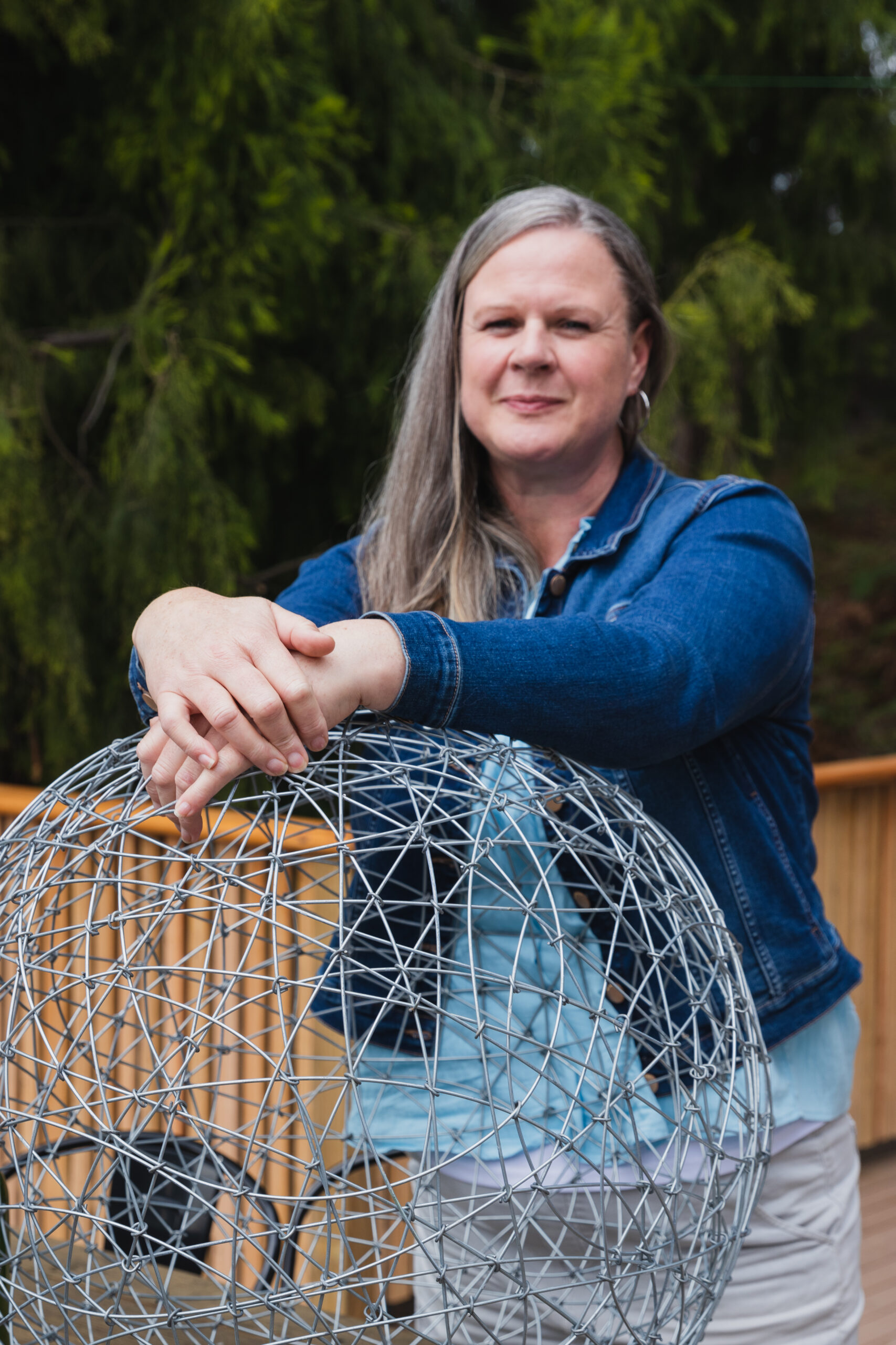 Kylie is tall, smiling and has long grey hair. Kylie is leaning on a wire sculpture. These wire sculptures are also sold at Laid Back Manor. There is a natural bush backdrop and timber handrail in the background.
